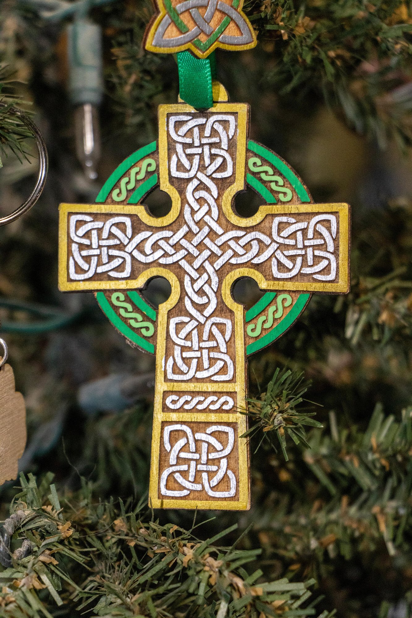 A wooden Celtic cross ornament with green and white knotwork, hanging on a Christmas tree