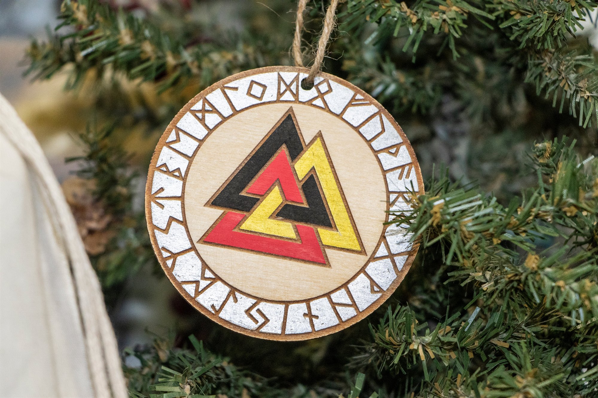 A round wooden ornament with a red, gold and black Valknut inside a ring of engraved runes, hanging on a Christmas tree
